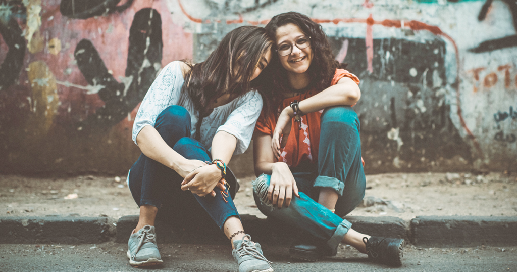 Two female friends smiling & sitting on the sidewalk 